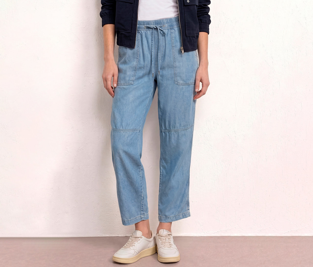 Une femme porte un jean bleu clair, des chaussures blanches et une veste bleu marine devant un mur blanc.
