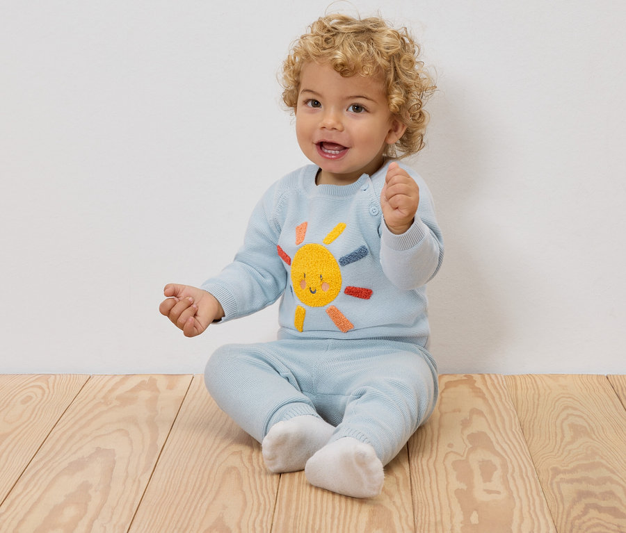 Un bébé souriant aux boucles blondes est assis sur un plancher en bois, portant un pull bleu clair avec un soleil brodé et un pantalon assorti.