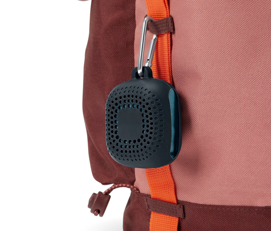 Un récipient perforé gris foncé avec un mousqueton est suspendu à une sangle orange d'un sac à dos marron rougeâtre.