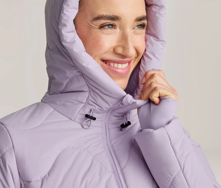 Femme souriante vêtue d'une veste matelassée violet clair à capuche, tenant le col.