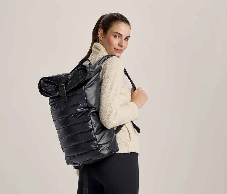 Femme portant un sac à dos noir matelassé brillant à enroulement et une veste polaire crème, regardant par-dessus son épaule.