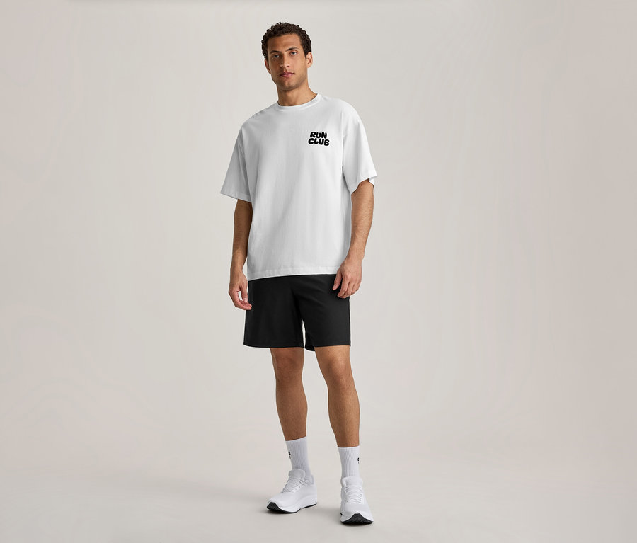 Un homme se tient de face, portant un T-shirt de sport blanc avec l'inscription noire « RUN CLUB » sur la poitrine, un short noir, des chaussettes blanches et des chaussures de sport blanches.