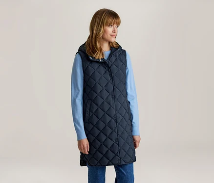 Femme aux cheveux bruns mi-longs avec une frange, portant une longue veste matelassée bleu marine à capuche sur une chemise à manches longues bleu clair et un jean bleu, regardant vers la droite.