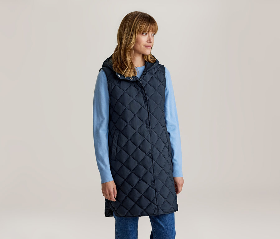 Femme aux cheveux bruns mi-longs avec une frange, portant une longue veste matelassée bleu marine à capuche sur une chemise à manches longues bleu clair et un jean bleu, regardant vers la droite.