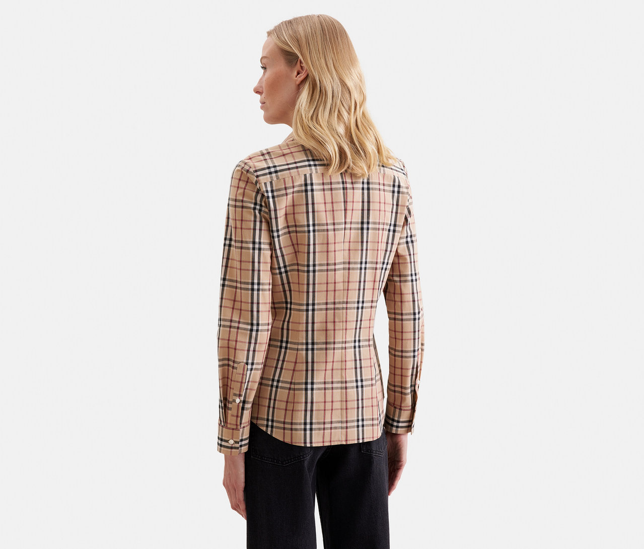 Femme aux longs cheveux blonds, vue de dos et légèrement tournée vers la droite, portant une chemise à manches longues beige à carreaux et un pantalon noir.