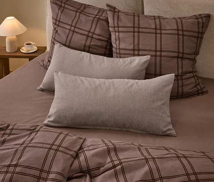 Un lit avec une couette à carreaux marron et deux grands oreillers assortis, deux oreillers rectangulaires gris clair unis et un drap-housse marron. Une lampe blanche et une tasse de café sont posées sur une table de chevet en bois.