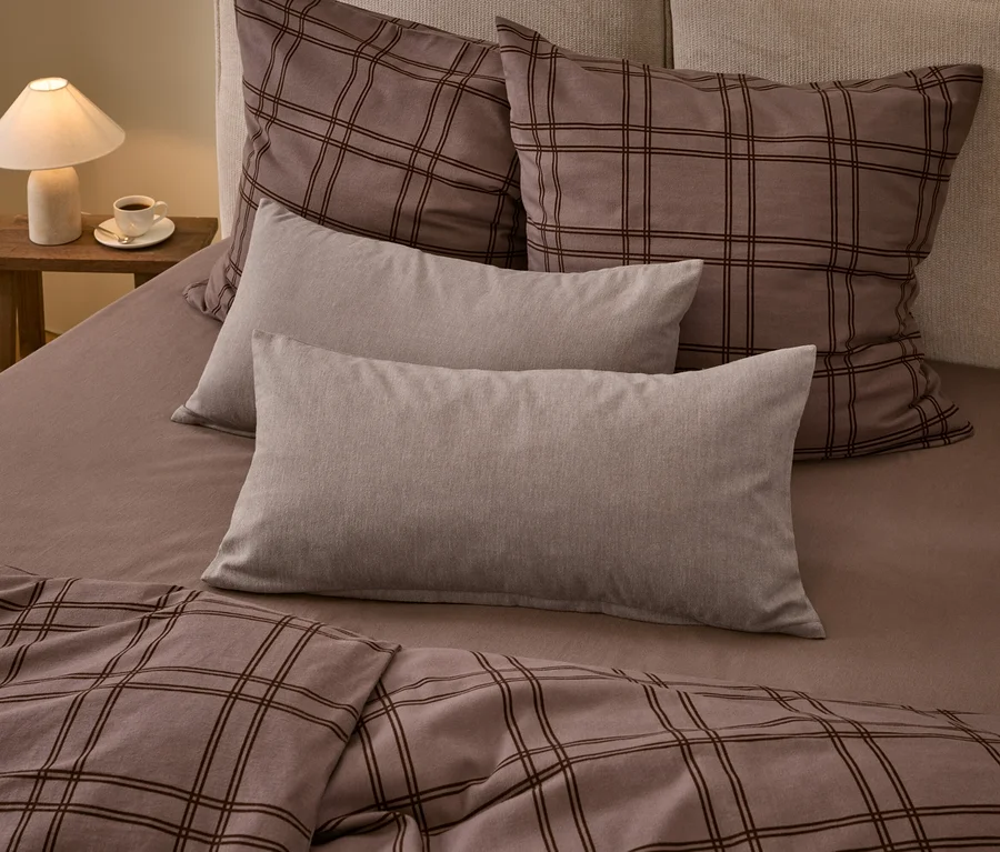 Un lit avec une couette à carreaux marron et deux grands oreillers assortis, deux oreillers rectangulaires gris clair unis et un drap-housse marron. Une lampe blanche et une tasse de café sont posées sur une table de chevet en bois.