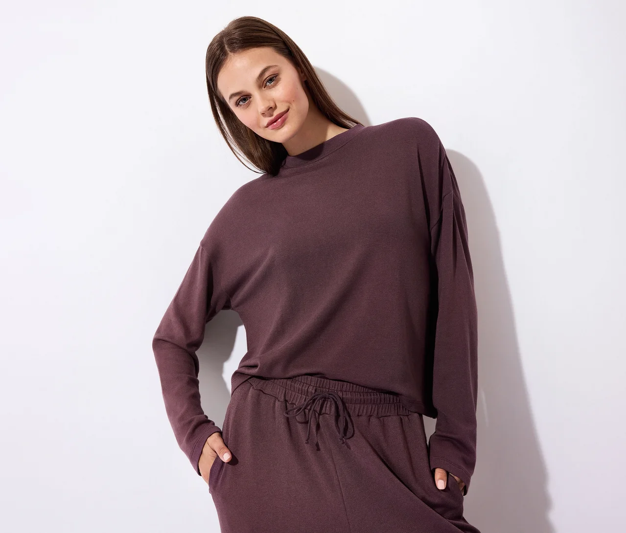 Femme portant un haut à manches longues et un pantalon de jogging assorti de couleur violet foncé, regardant l'appareil photo avec une main dans sa poche.