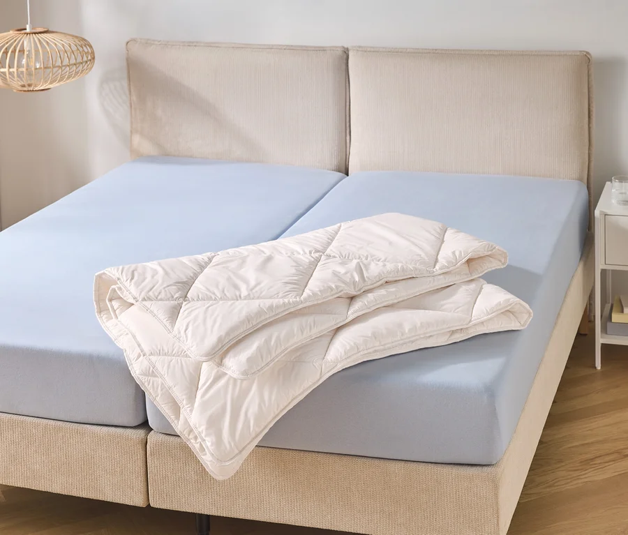 Un lit double avec une tête de lit beige et deux draps-housses bleu clair. Une couette matelassée crème pliée repose sur le côté droit du lit.