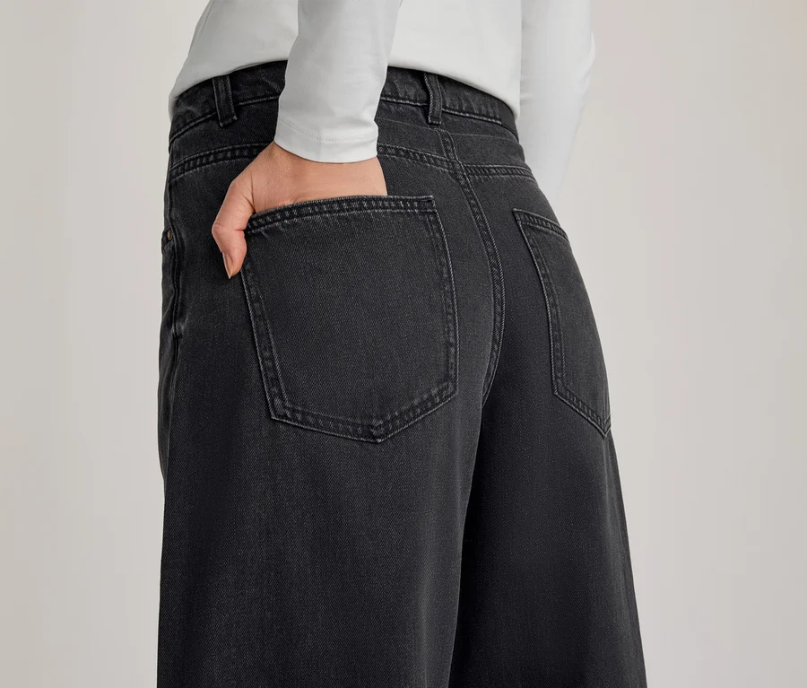 Personne de dos, portant un jean gris foncé à jambes larges et un haut blanc à manches longues, une main dans la poche arrière du jean.