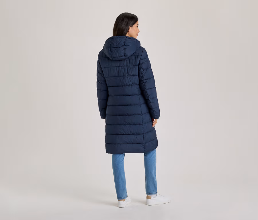 Femme debout de dos, portant un long manteau matelassé bleu marine à capuche, un jean bleu et des baskets blanches.