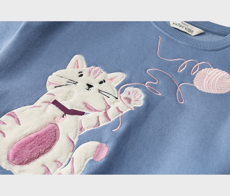 Gros plan d'un sweat-shirt bleu avec une application de chat blanc à rayures roses et ventre rose duveteux, tendant la patte vers un fil et une pelote de laine roses brodés.