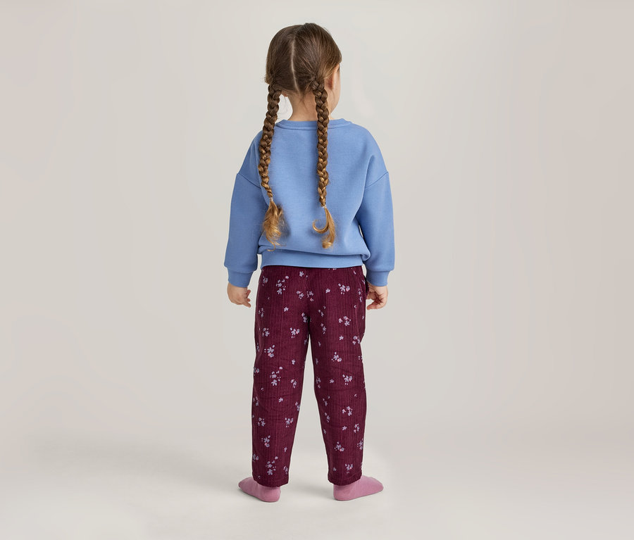 Enfant de dos, portant un pull bleu et un pantalon bordeaux à motifs, avec deux tresses.