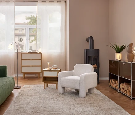 Un salon confortable avec un canapé vert, un fauteuil clair et un poêle à bois noir. Une commode et une table d'appoint sont près de la fenêtre avec des rideaux. À droite, une étagère marron avec des bûches et une lampe de table.
