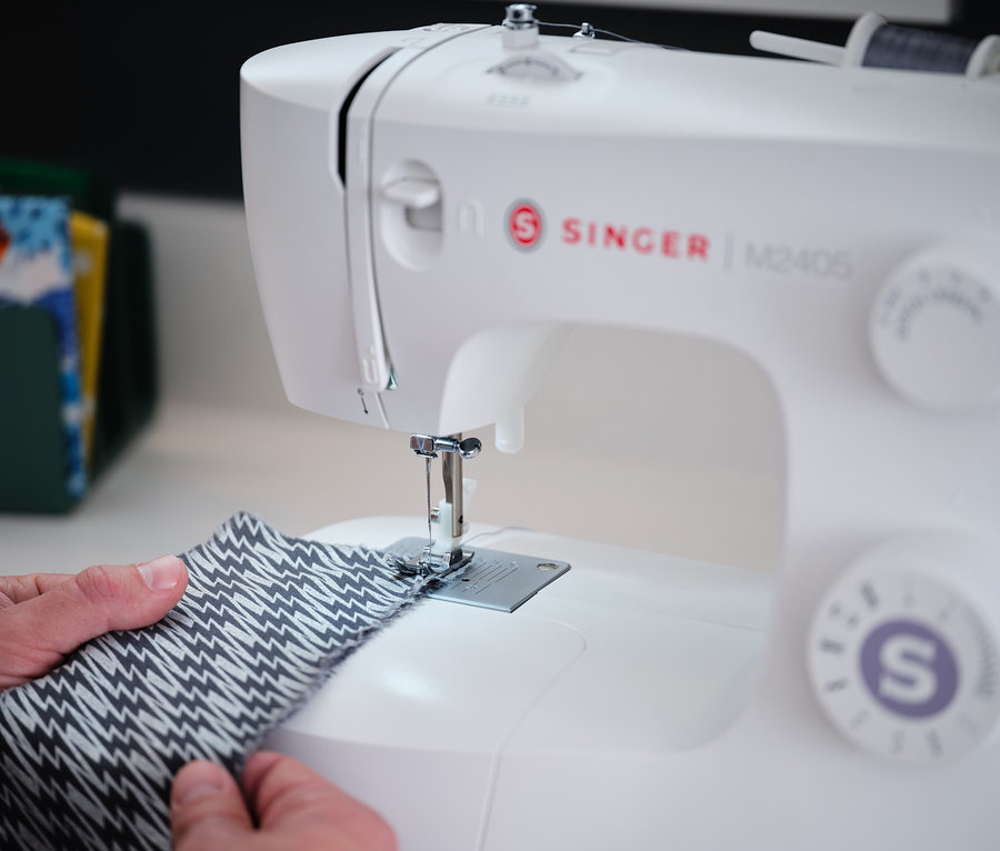 Les mains guident un morceau de tissu à motifs sous l'aiguille de la machine à coudre Singer «
M2405
».