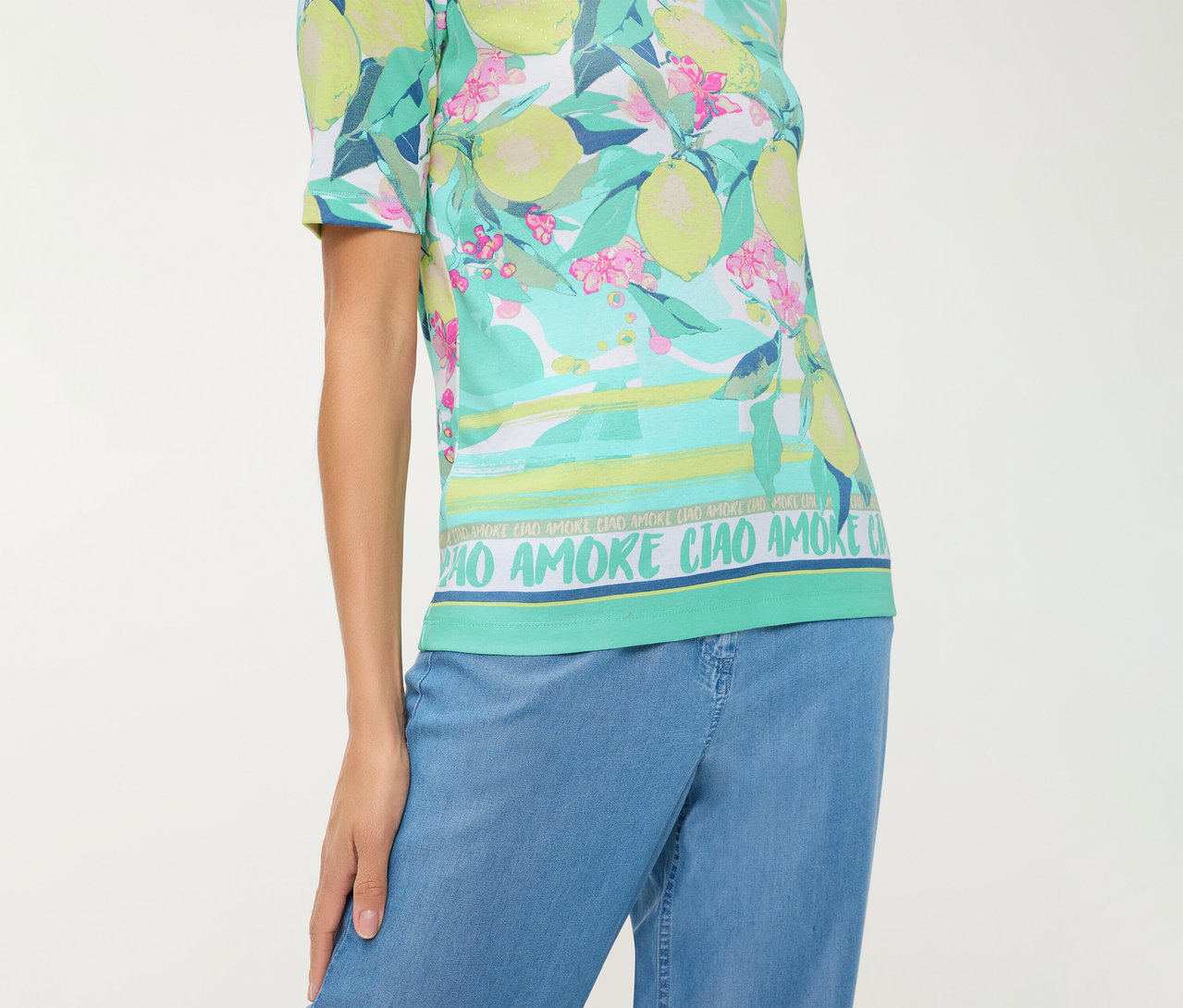 Personne portant un T-shirt Olsen à motif citrons et fleurs, rayures horizontales et texte "CIAO AMORE", avec un pantalon bleu.