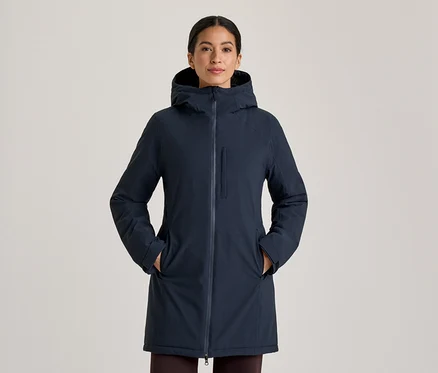 Femme portant un manteau à capuche bleu foncé, debout les mains dans les poches du manteau. Le manteau a une fermeture éclair intégrale et une poche poitrine verticale zippée.