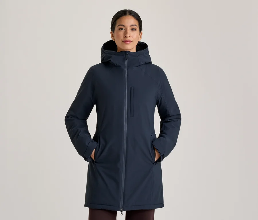 Femme portant un manteau à capuche bleu foncé, debout les mains dans les poches du manteau. Le manteau a une fermeture éclair intégrale et une poche poitrine verticale zippée.