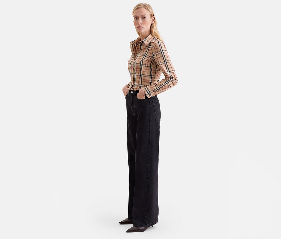 Femme blonde debout, les mains dans les poches, portant une chemise à manches longues à carreaux beige, un jean noir large et des talons noirs pointus.