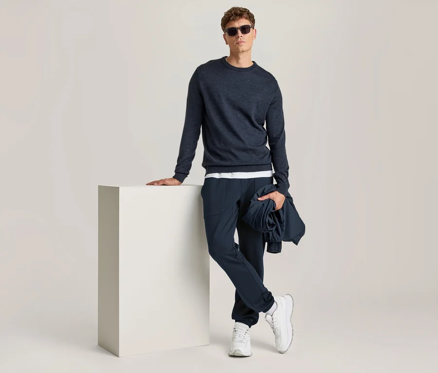 Un homme debout, appuyé sur un cube blanc, porte un haut à manches longues bleu foncé, un pantalon bleu foncé, des baskets blanches et des lunettes de soleil, il tient une veste bleu foncé roulée.