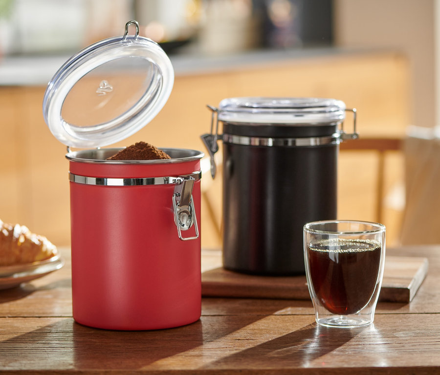 Sur une table en bois se trouvent une boîte à arômes rouge ouverte et une noire fermée à côté d'un verre de café et d'une assiette avec un croissant.