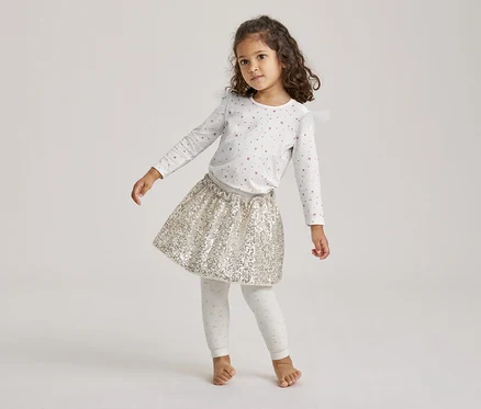 Une petite fille aux cheveux bouclés se tient pieds nus, vêtue d'un t-shirt blanc à manches longues avec des étoiles roses, d'une jupe dorée à paillettes et de leggings à pois.