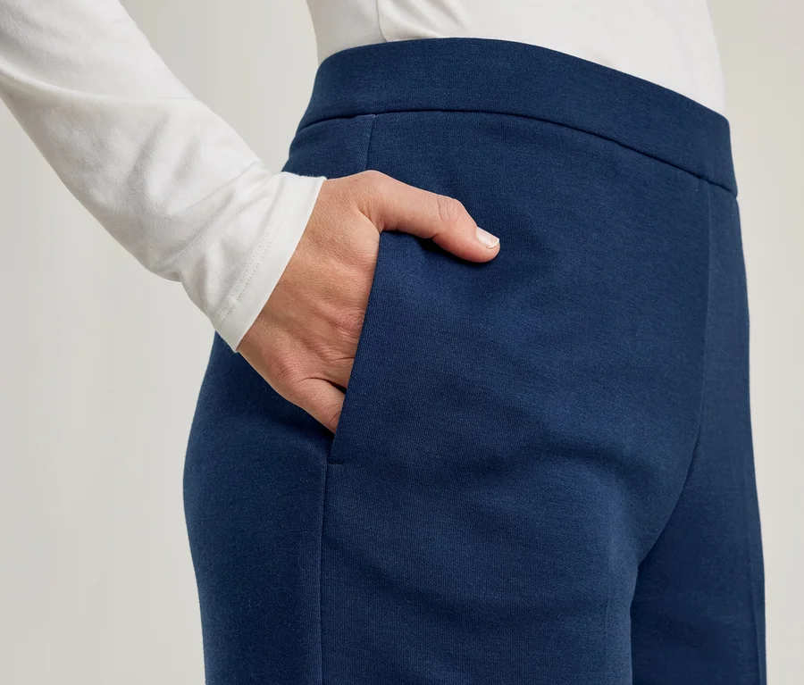 Une personne portant un haut blanc à manches longues et un pantalon bleu foncé, avec sa main droite placée dans la poche du pantalon.