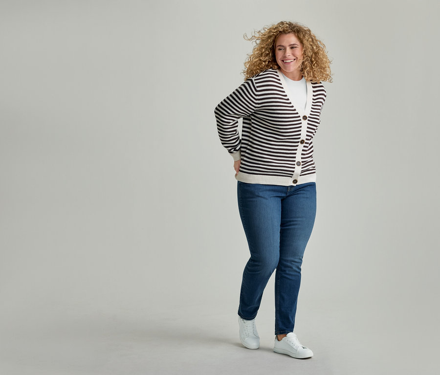Femme souriante aux cheveux bouclés portant un cardigan rayé, un t-shirt blanc, un jean bleu et des baskets blanches, les mains derrière le dos.