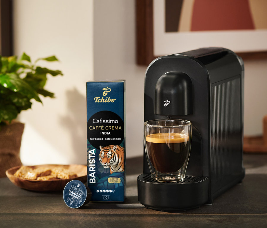 Machine à café noire Tchibo Cafissimo avec un verre de café, à côté une boîte de capsules de café Tchibo Cafissimo Caffè Crema India et une capsule individuelle.