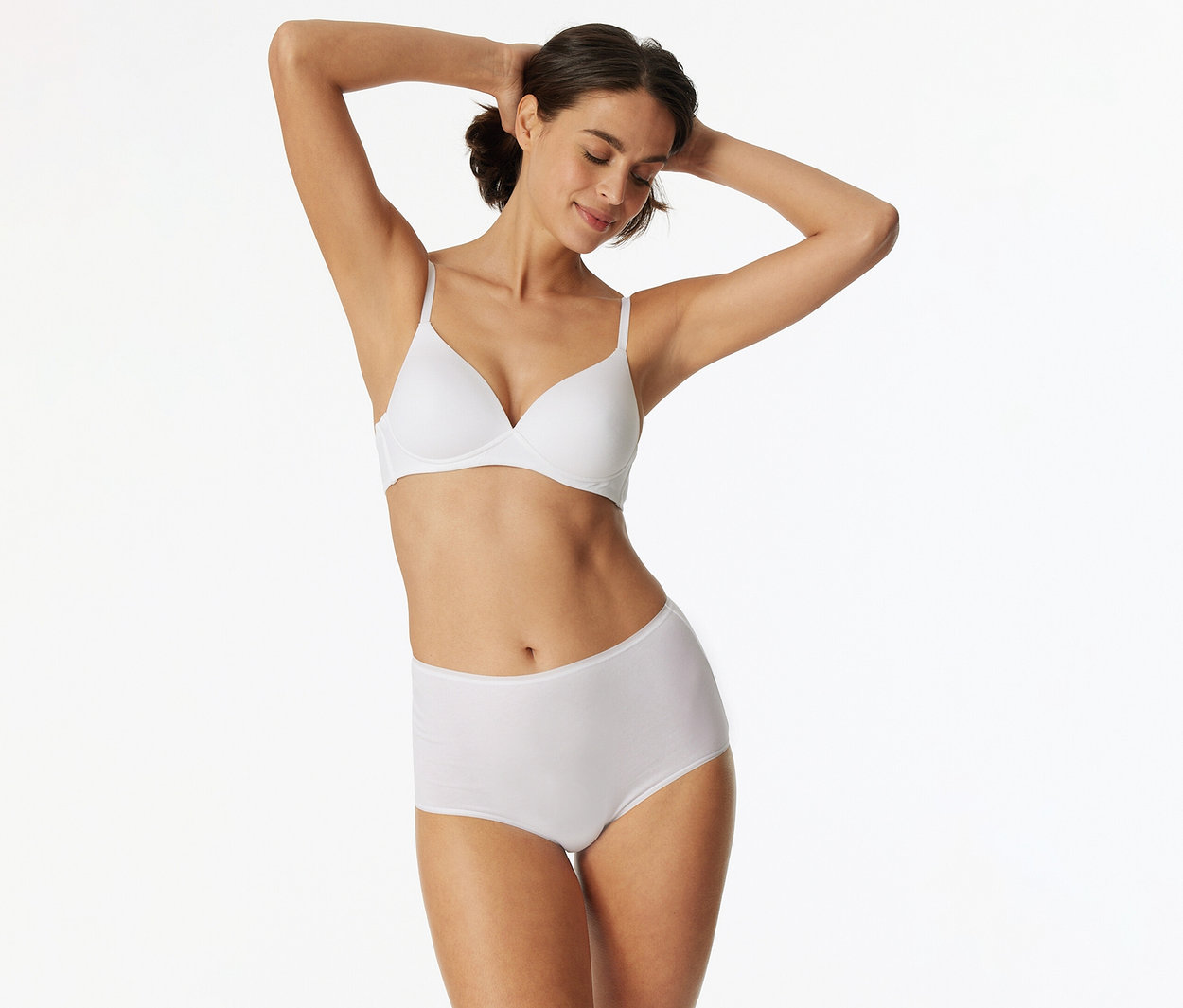 Femme aux cheveux foncés, portant un soutien-gorge blanc et une culotte taille haute blanche, les bras levés derrière la tête, les yeux fermés et souriante.