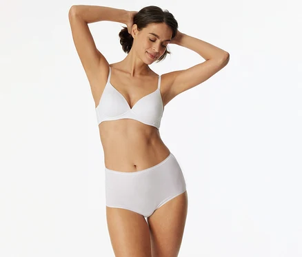 Femme aux cheveux foncés, portant un soutien-gorge blanc et une culotte taille haute blanche, les bras levés derrière la tête, les yeux fermés et souriante.