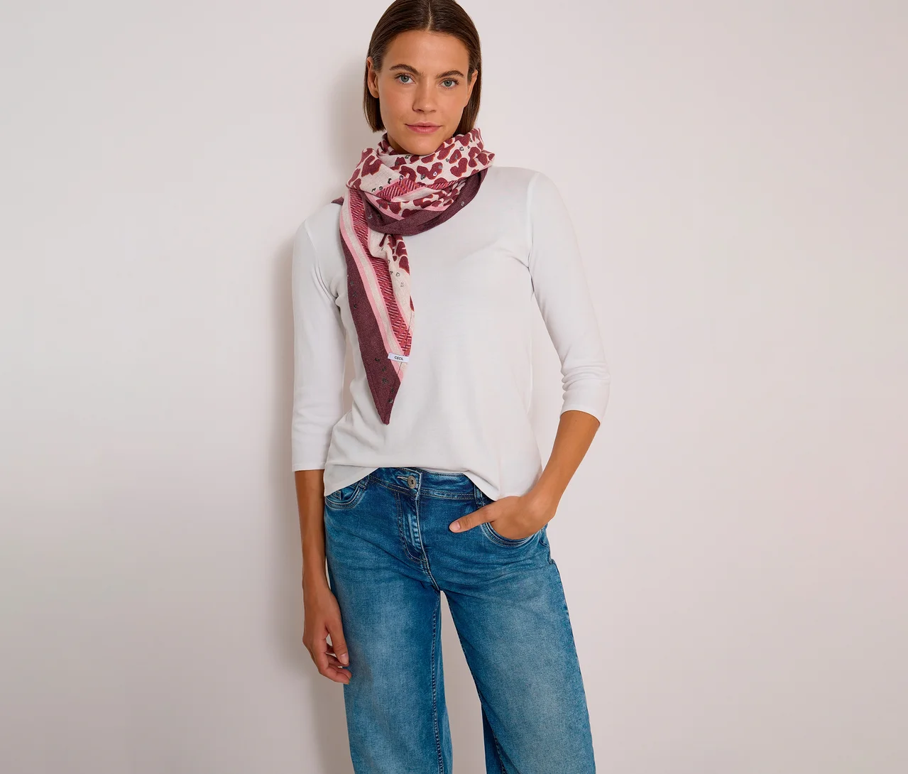 Une femme porte un haut blanc à manches longues, un jean et un foulard triangulaire CECIL à motifs dans des tons de rose et de bordeaux.