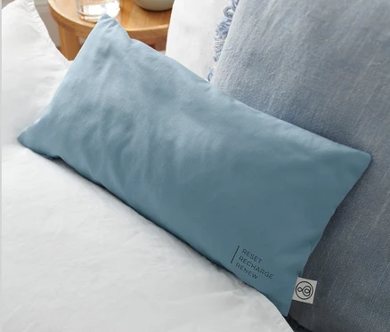 Un coussin bleu garni de copeaux de pin cembro, env. 40 x 20 cm, est posé sur du linge de lit blanc à côté d'un coussin décoratif bleu.