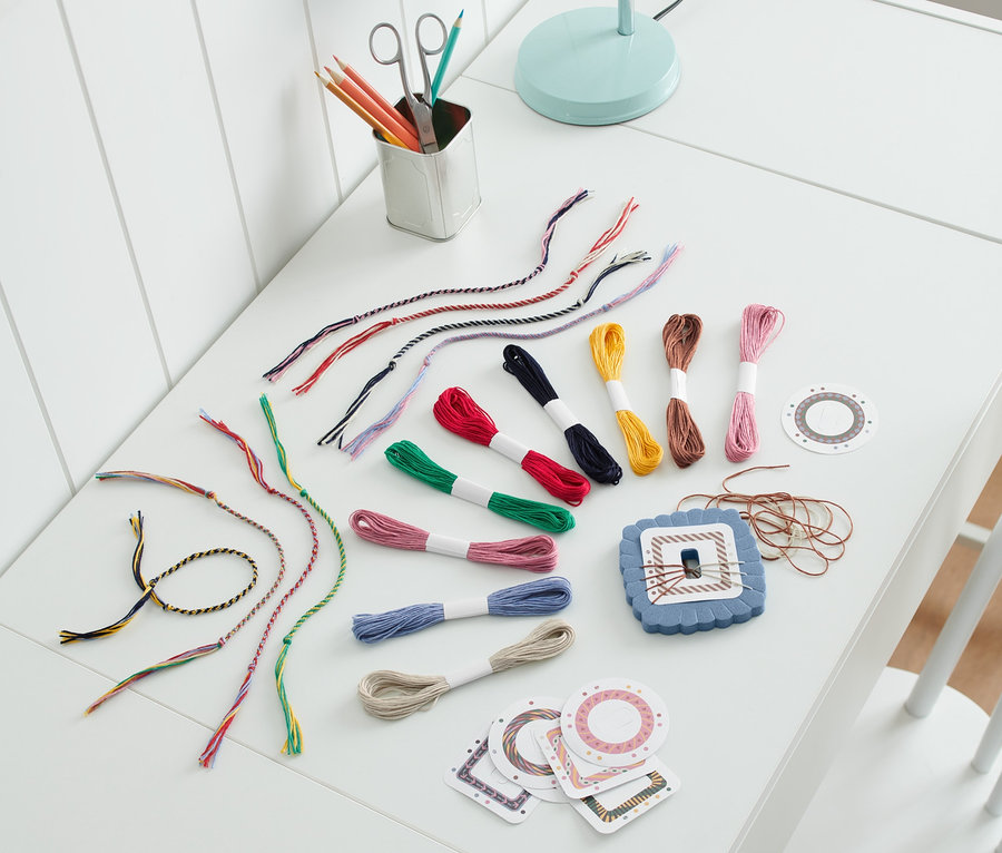 Sur une table blanche se trouvent un kit de bricolage « bracelets brésiliens » ainsi que des fils de coton colorés, des ciseaux et des crayons de couleur.