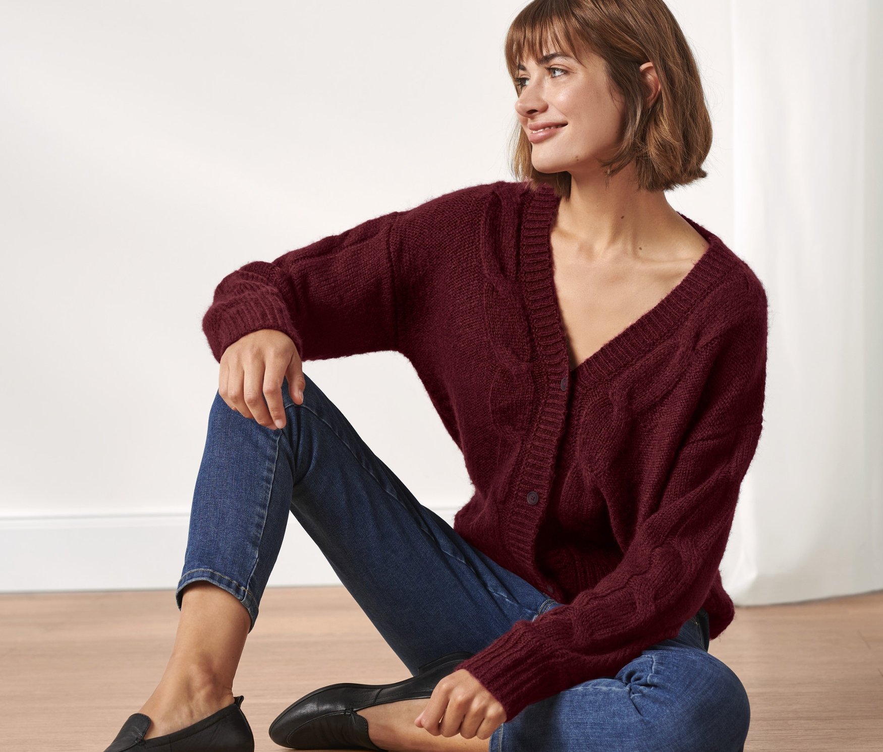 Une femme est assise par terre et porte un cardigan avec torsades, un jean et des mocassins noirs.