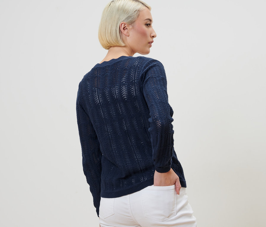 Une femme, vue de dos, porte une jaquette en maille à motif ajouré et un jean blanc.