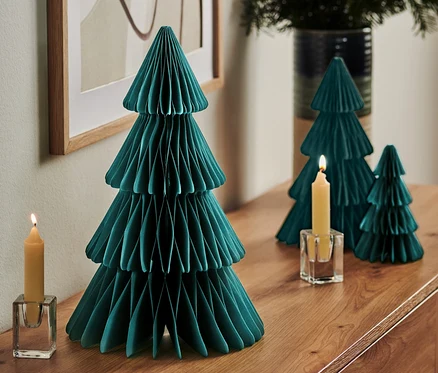 Sur une commode se trouvent un grand arbre en papier pliable, deux petits arbres, une bougie allumée dans un bougeoir en verre et un vase avec des branches.