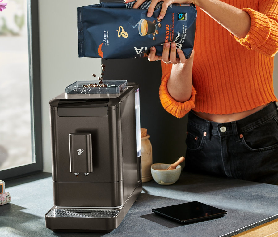Une femme portant des lunettes verse des grains de café dans une machine à café entièrement automatique « Esperto2 Caffè », Granite Black.