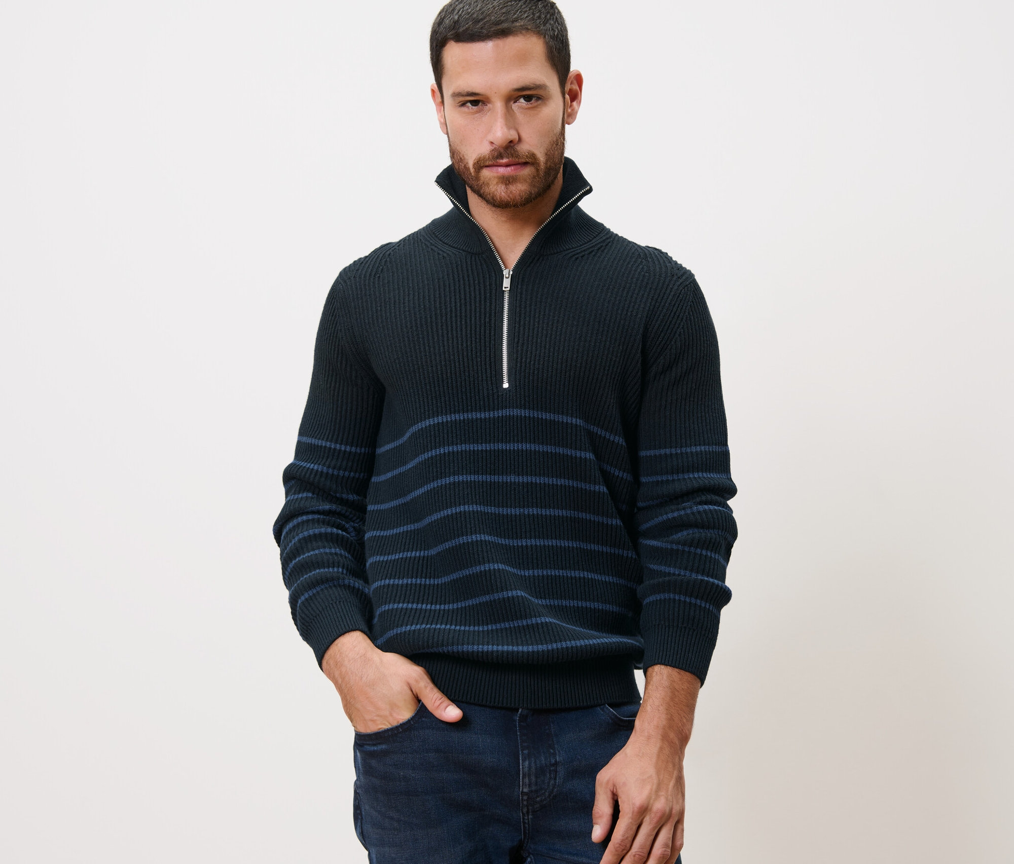 Un homme pose en pull camionneur, la main dans la poche de son jean.