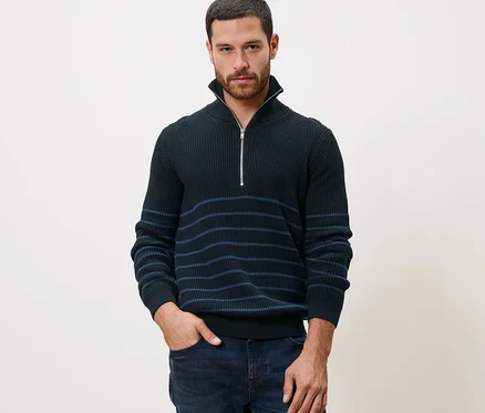 Un homme pose en pull camionneur, la main dans la poche de son jean.