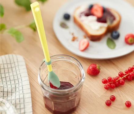 Cuillère spéciale en silicone jaune dans un pot de confiture à côté d'une assiette de pain grillé aux fruits.