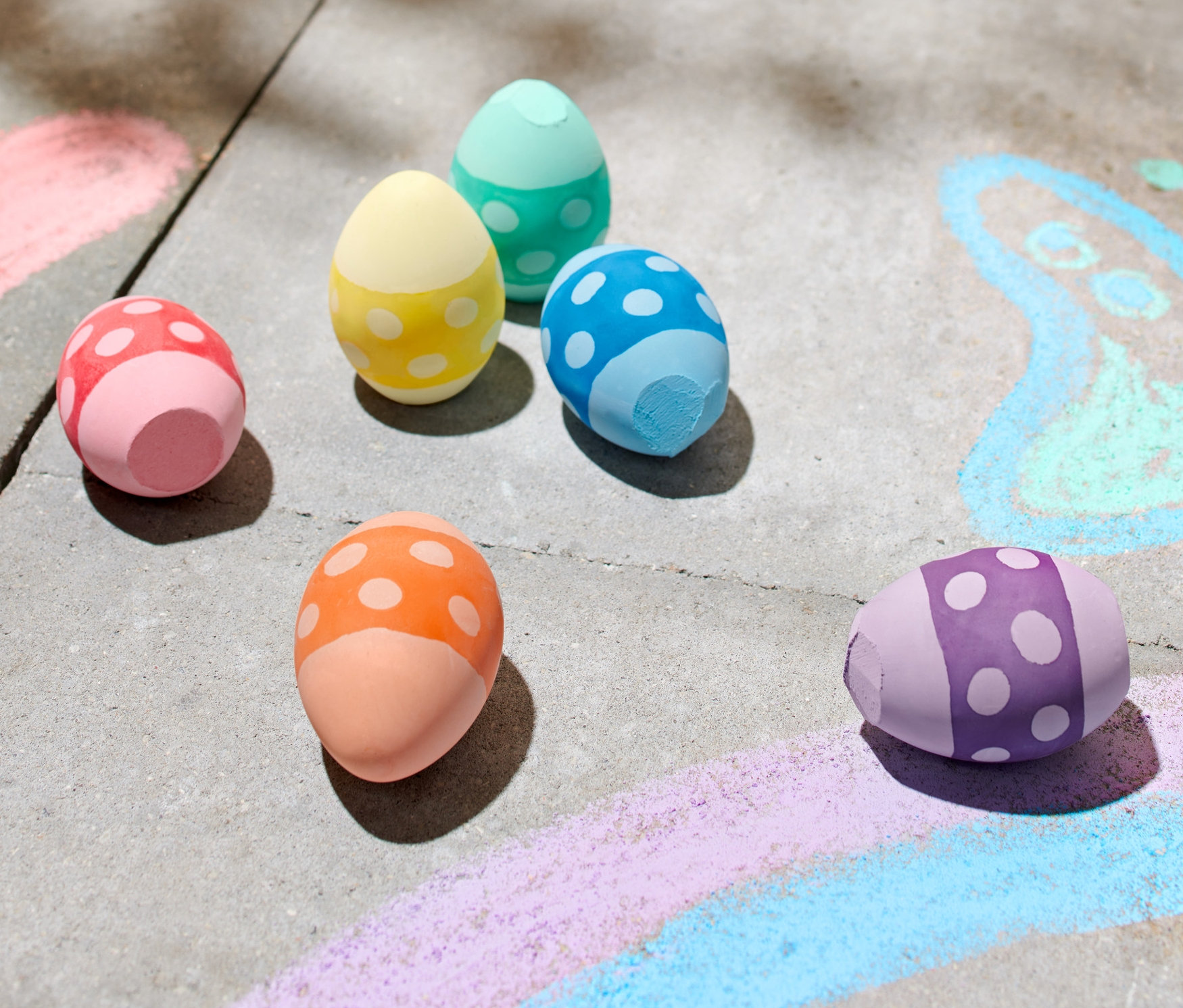 Six craies d'extérieur colorées en forme d'œufs sont posées sur un trottoir à côté de dessins à la craie.