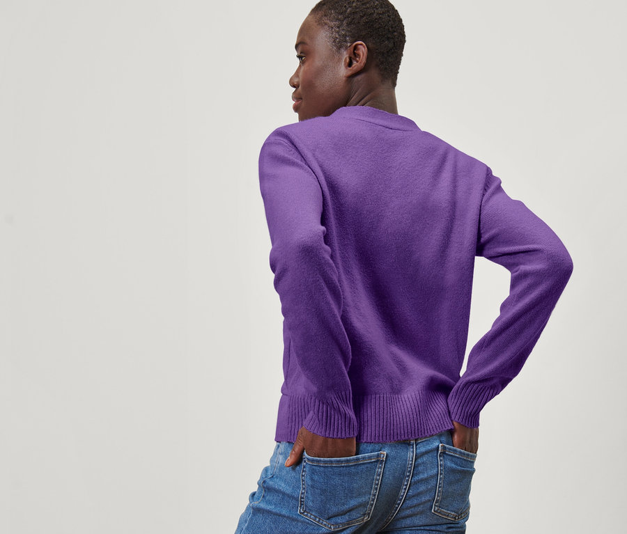 Une femme pose, dos à la caméra, les mains dans les poches de son jean. Elle porte une jaquette en maille violette avec de la laine.