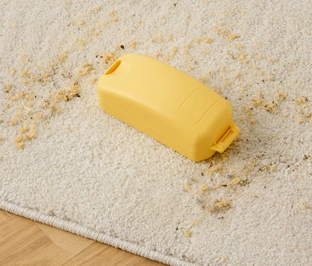 Un ramasse-miettes jaune est posé sur un tapis beige avec des miettes éparpillées.