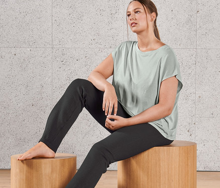 Une femme est assise sur un tabouret en bois et porte un t-shirt de yoga et un pantalon de sport évasé sans coutures, avec fente.