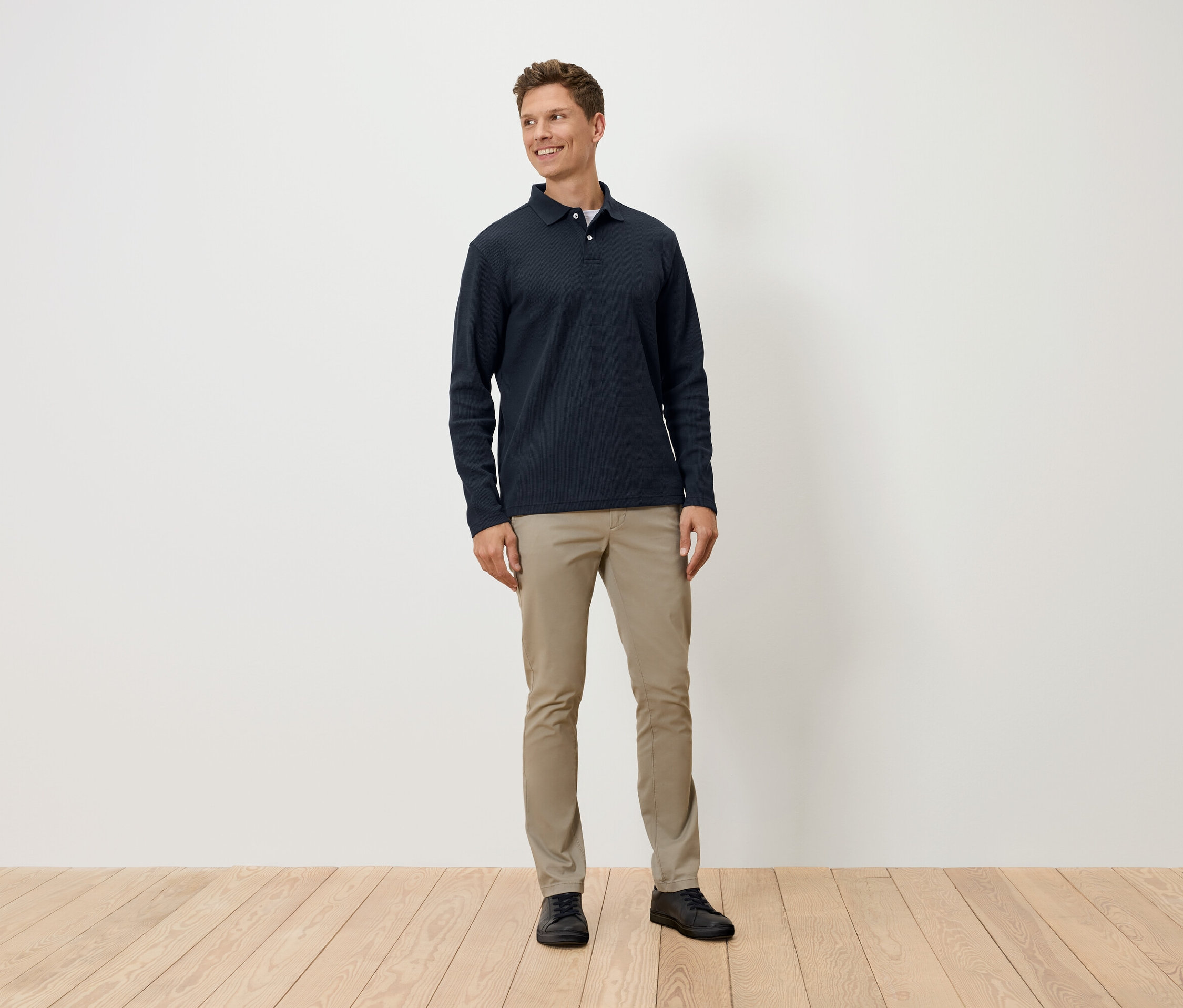 Un homme debout porte un polo à manches longues bleu marine, un pantalon beige et des chaussures noires.