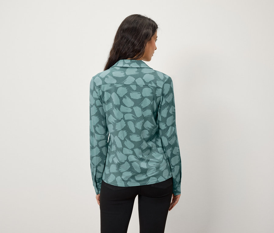 Vue de dos d'une femme aux longs cheveux foncés portant une chemise bleu-vert à motifs et un pantalon noir.