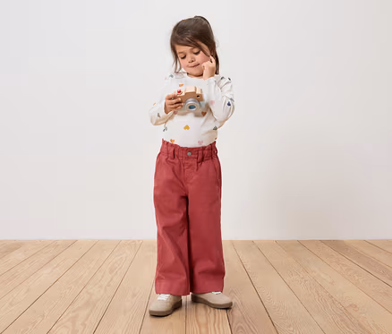 Une petite fille tient un appareil photo en bois et le regarde. Elle porte un pantalon et un t-shirt à manches longues.