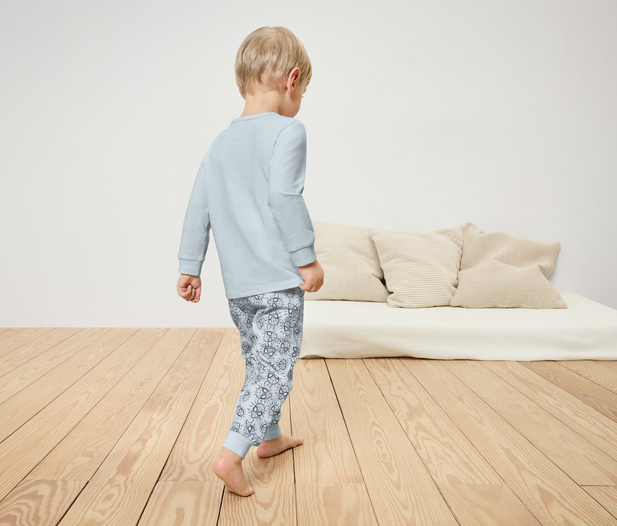 Un garçon en pyjama marche sur un plancher en bois. Un lit avec des oreillers est visible en arrière-plan.