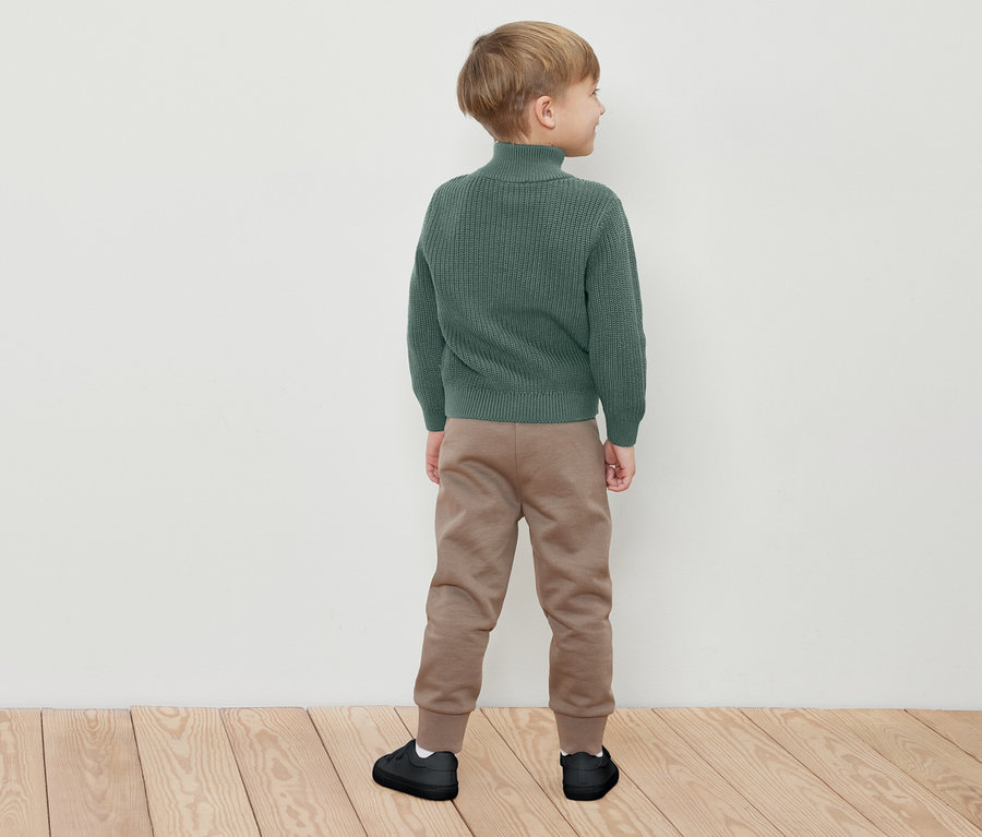 Un garçon se tient dos à la caméra, portant un pull à col roulé vert, un pantalon de jogging marron et des baskets noires.